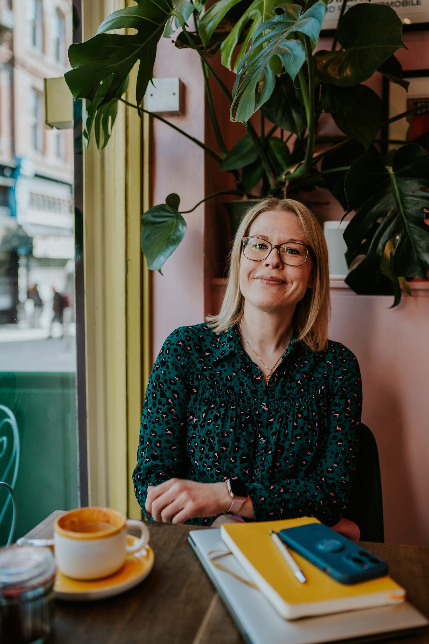 Woman sat in cafe, with a Monstera plant behind her, sat at a table with her mobile phone, note book and laptop stacked in front of her and a cappuccino. 