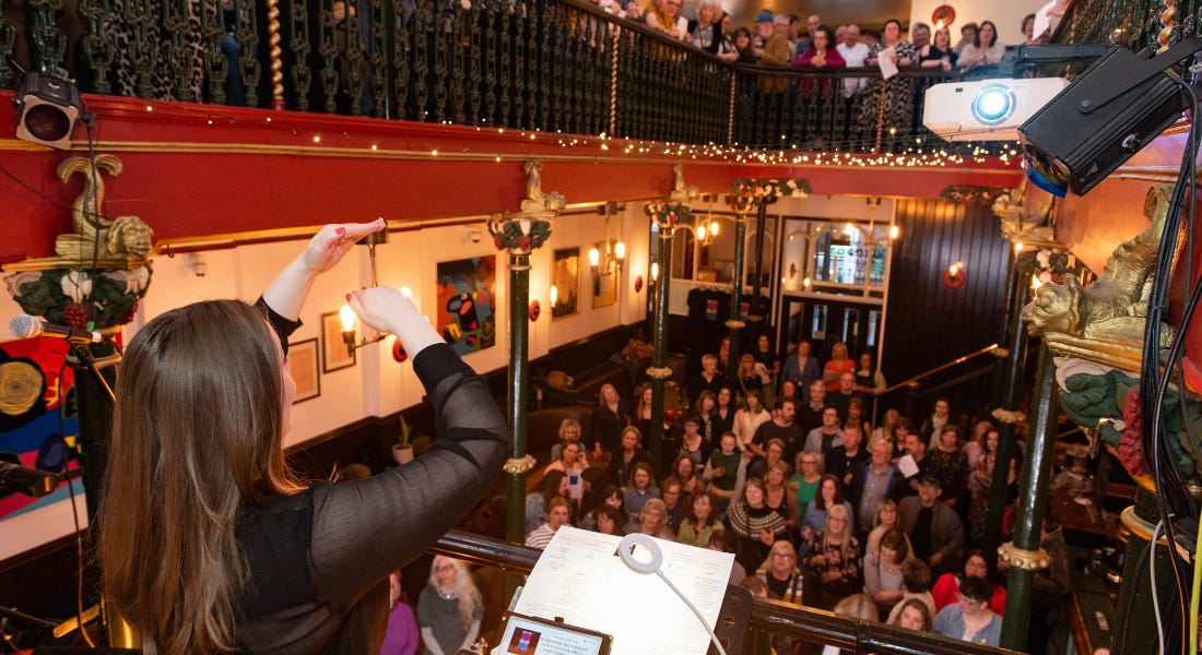 A music hall with a woman at the front conducting and a crowd of people in front of her singing
