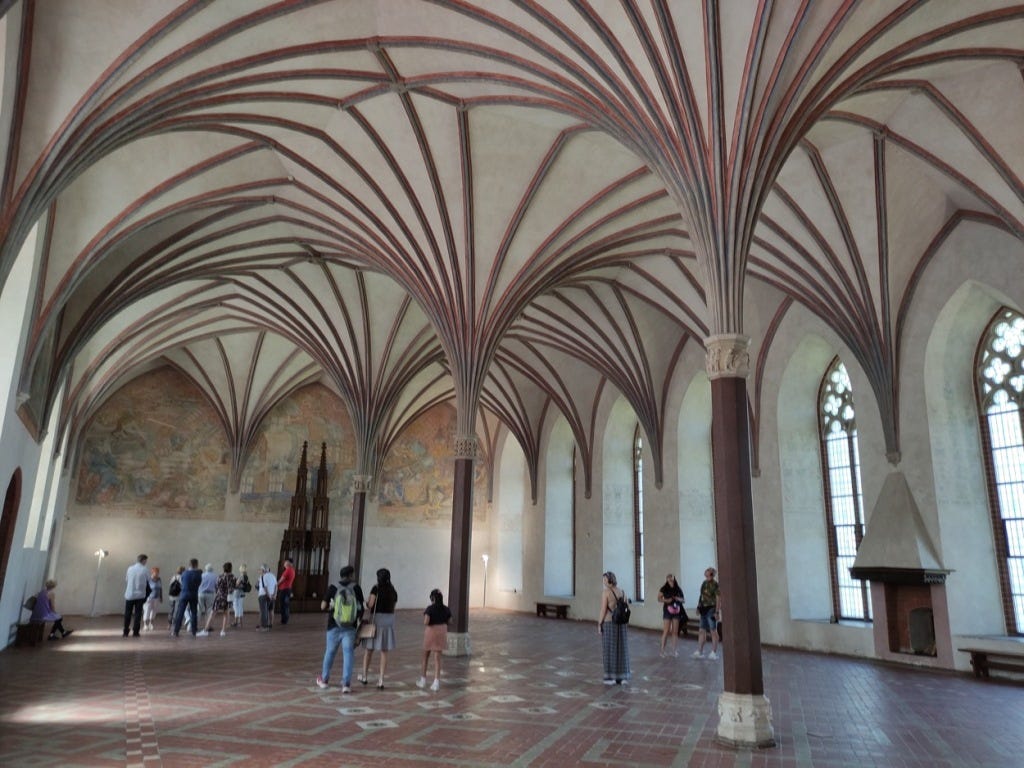 A very large Great Hall, with a small fireplace to one side. Pillars along the middle of the hall hold up curved arches that spiral out across the ceiling