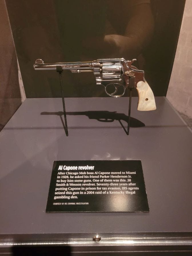 Numa redoma de vidro está apoiada em destaque um revólver com cabo branco e amadeirado e o corpo metálico. A arma pertenceu ao mafioso Al Capone.