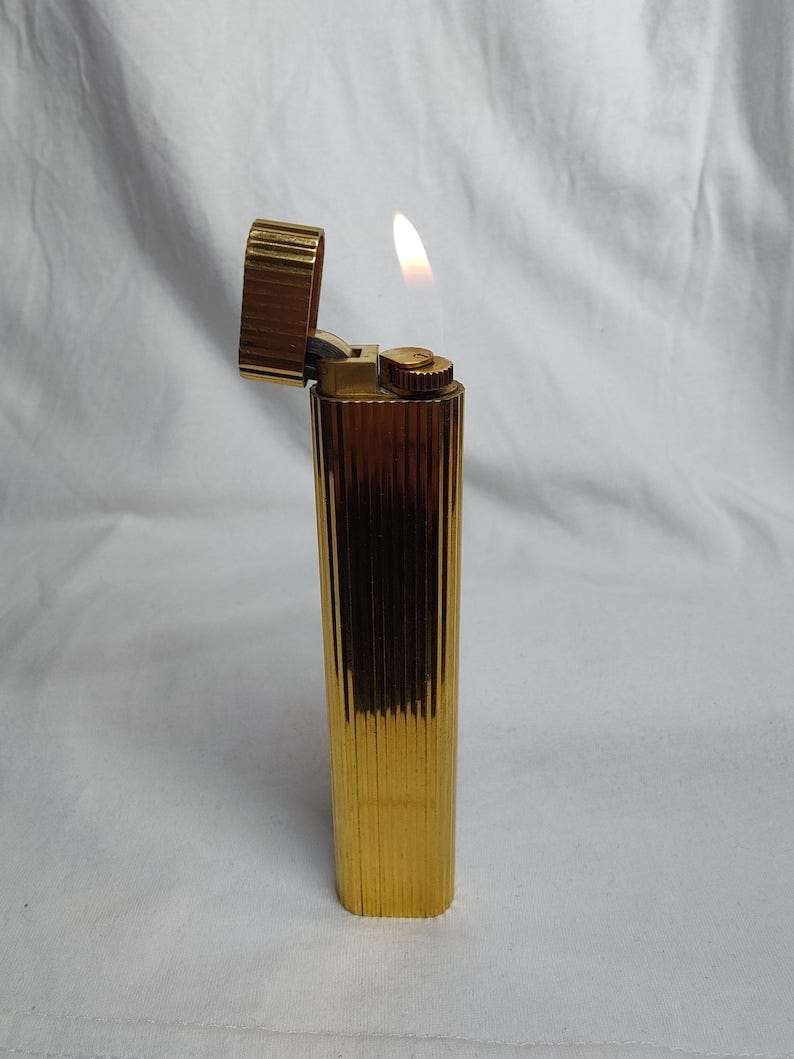 May include: A gold-coloured lighter with a vertical ribbed design. The lighter is open, and a flame is lit. The lighter has a rectangular shape with rounded edges. The background is a white fabric.