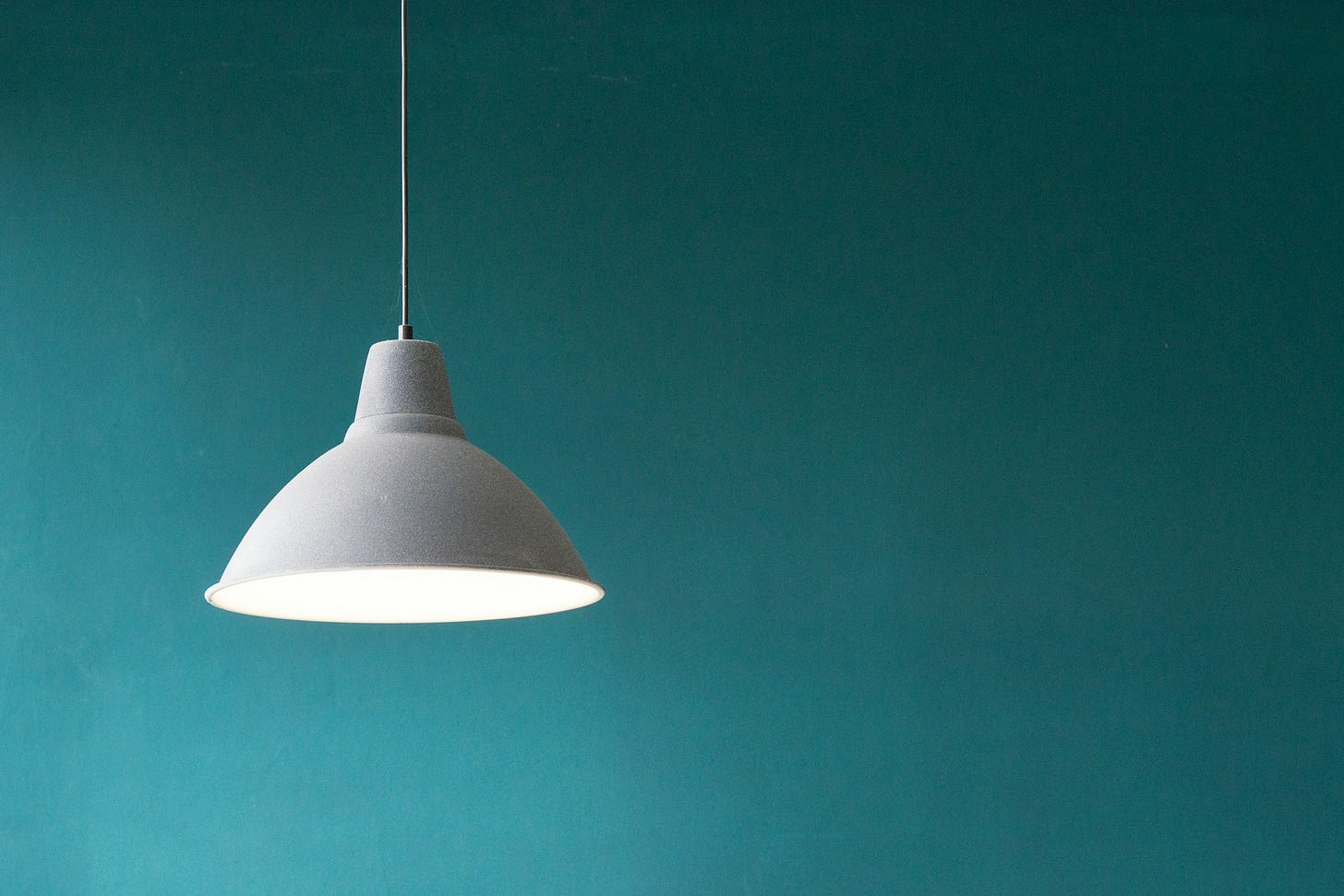 Sfondo verde acqua con sfumatura che si schiarisce dall'alto verso il basso. In primo piano, pende a sinistra un lampadario con filo e plafoniera bianche. La luce all'interno è accesa. Sfondo verde acqua con sfumatura che si schiarisce dall'alto verso il basso. In primo piano, pende a sinistra un lampadario con filo e plafoniera bianche. La luce all'interno è accesa.
