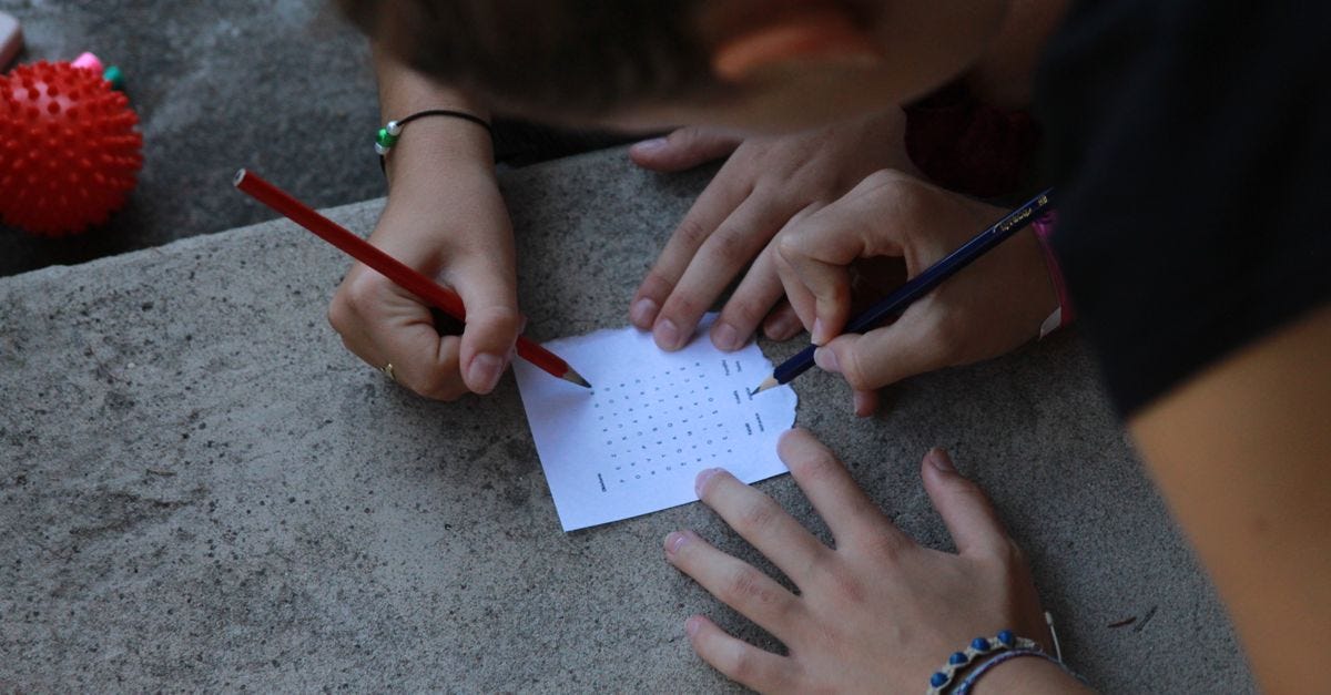 Close-up of hands solving a word puzzle outdoors using pencils on paper.