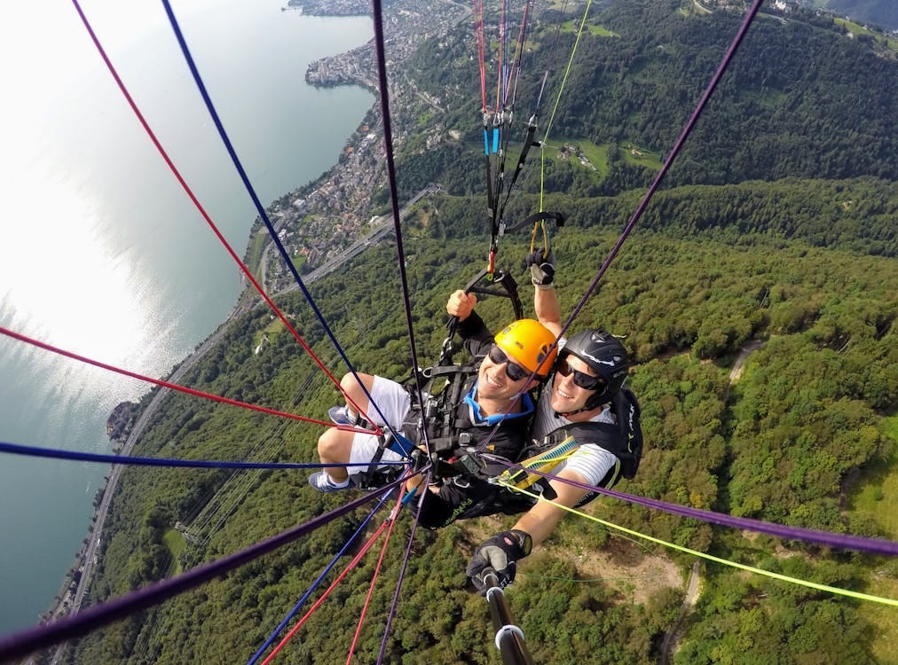 Free Two adults paragliding over a scenic landscape with forest and lake views, captured from above. Stock Photo
