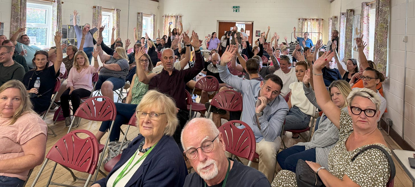 A large group of people gathered in a community hall, seated on red folding chairs arranged in rows. Several individuals have raised their hands, suggesting a vote or active participation. The room has white walls, floral curtains, and a wooden floor, creating a warm, informal setting.