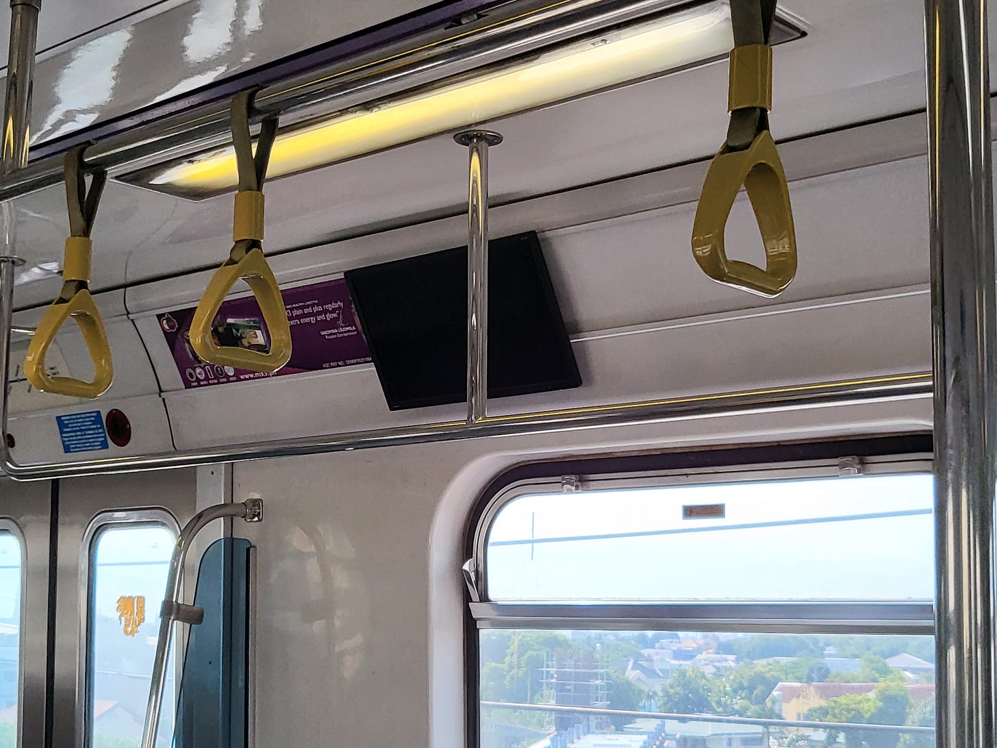 An interior shot of an LRTA 2000 Class train car, with a focus on a single monitor with its screen turned off. Nearby are slide-on advertising, handrails, and the safety straps