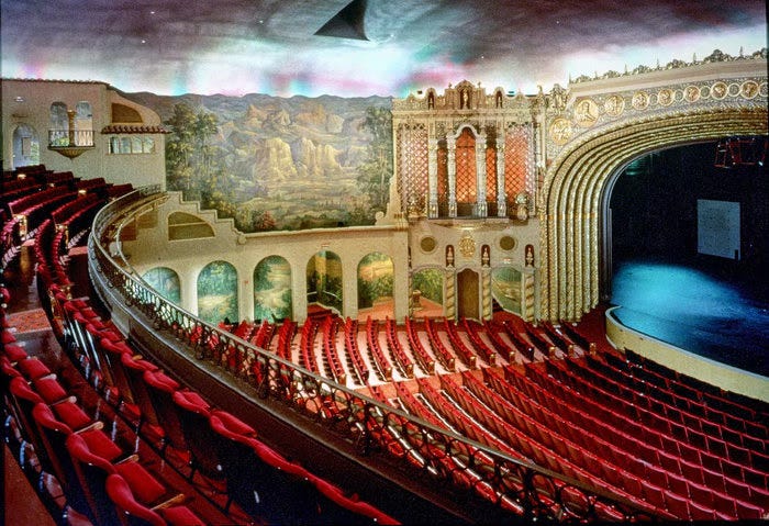 An opulent theater with red velvet seats, gilt columns, and desert-themed murals