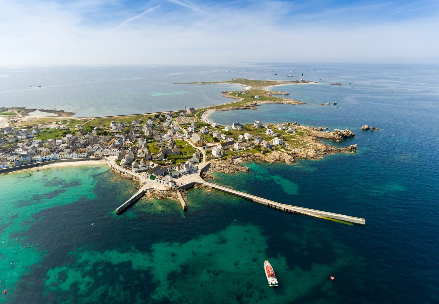 Aerial view of a coastal village with stone houses, a long pier, clear turquoise water, and a red boat near the shore, surrounded by small islands and the open sea.