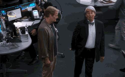Gif dalla serie tv "The Newsroom". Due uomini in un ufficio moderno e caotico. Uno indossa abiti casual marroni, l'altro un completo scuro con papillon grigio e ha un'espressione buffa o scettica. Sullo sfondo si vedono scrivanie con computer e persone in movimento.