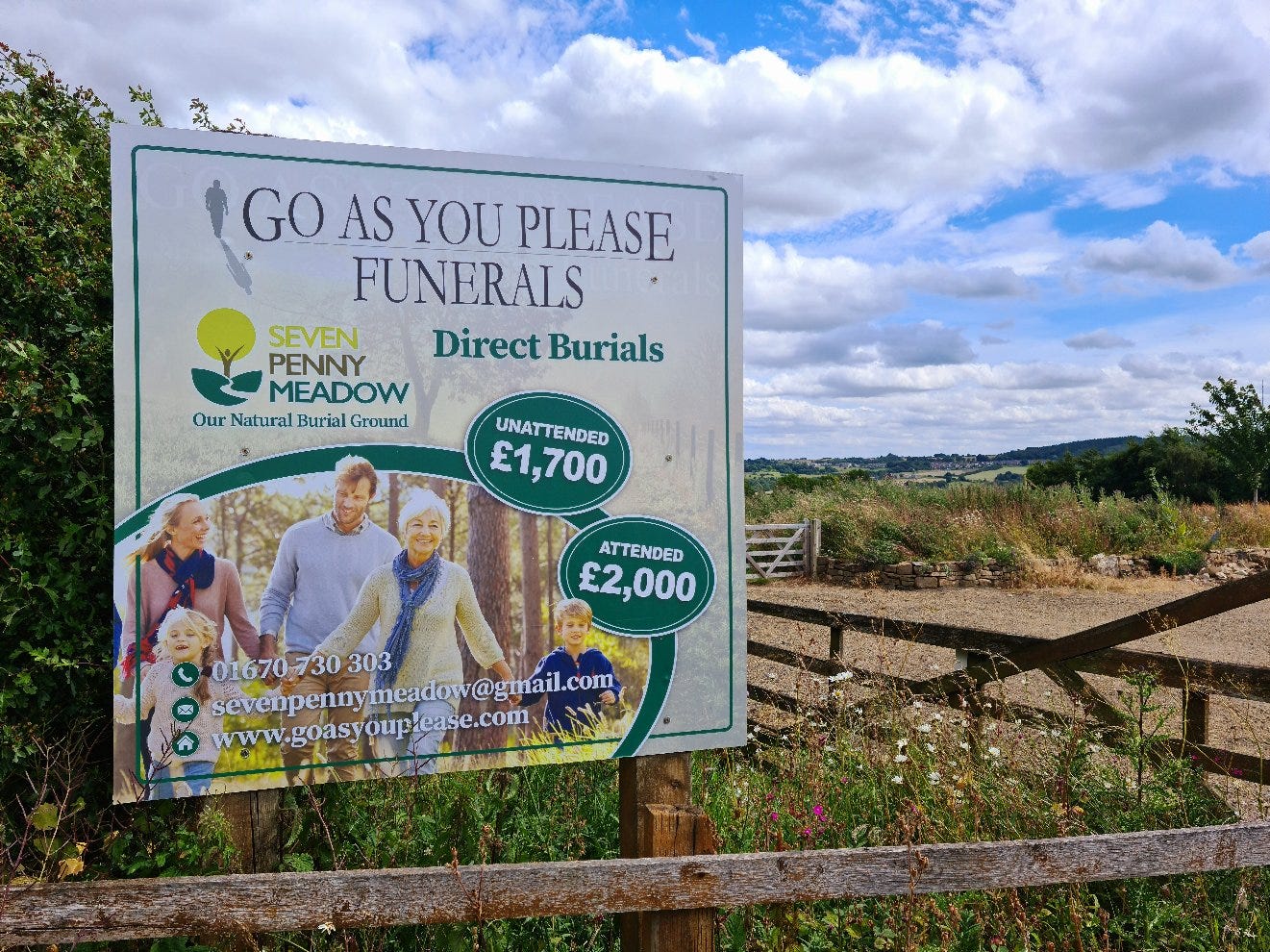 Entrance to a hilltop meadow that is a natural burial ground. Expansive blue sky with white clouds. Sign in the foreground that outlines the costs of direct burials at Seven Penny Meadow. Entrance to a hilltop meadow that is a natural burial ground. Expansive blue sky with white clouds. Sign in the foreground that outlines the costs of direct burials at Seven Penny Meadow.
