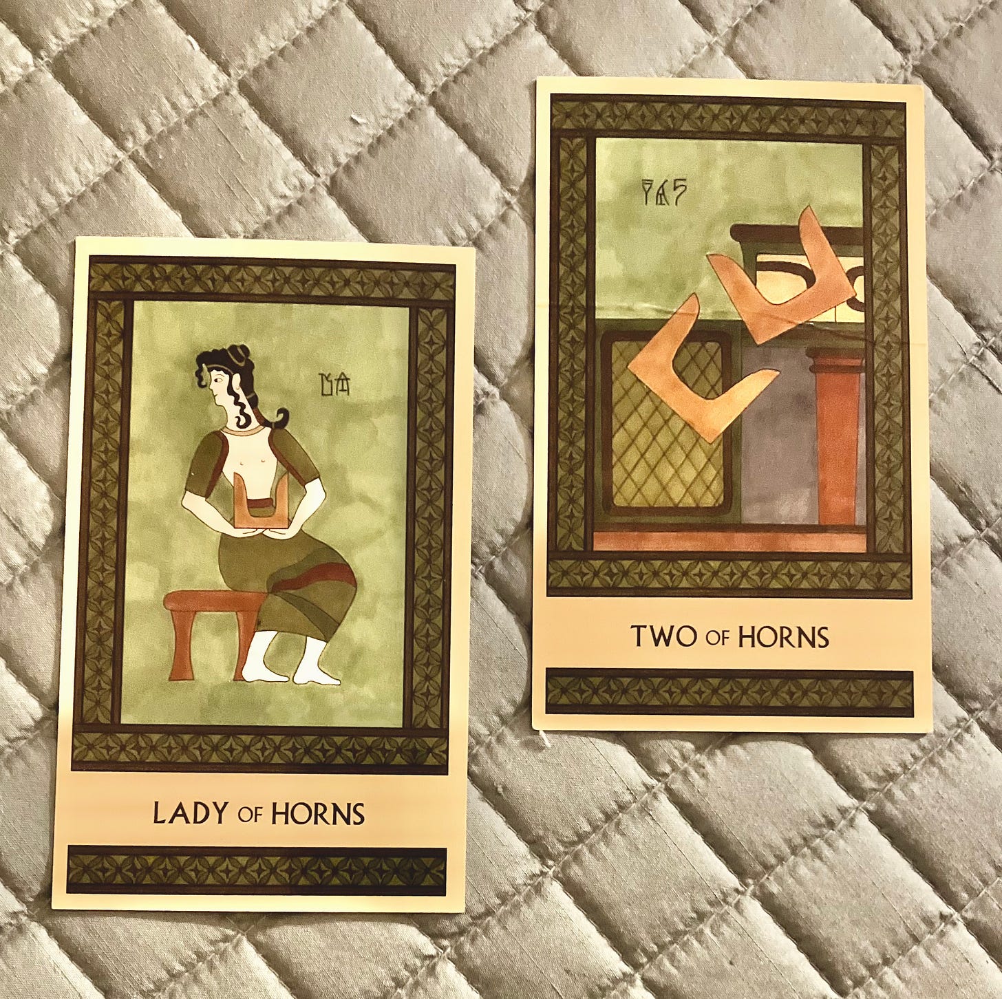 Two Minoan Tarot cards side by side on a silver quilted fabric surface. Both are painted in shades of green and brown. The Lady of Horns shows a Minoan woman seated on a bench, holding a pair of sacred horns. Her body faces right but her head is turned back to the left. The Two of Horns shows two pairs of sacred horns in midair as they tumble off a shrine.