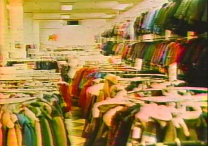 A faded colour photo of racks of clothes in a department store. The store is free of people.