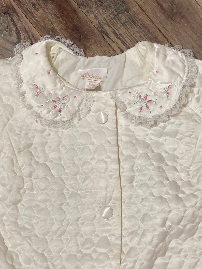 May include: A white quilted robe with a lace collar and pink floral embroidery. The robe has a button closure and a label that reads 'Bartholomew'.