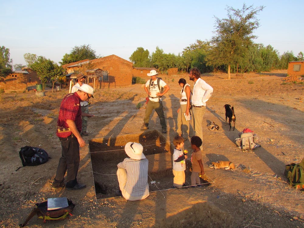 A group of people and their children surround an open archaeological dig site behind which stand a three buildings and some trees. A group of people and their children surround an open archaeological dig site behind which stand a three buildings and some trees.