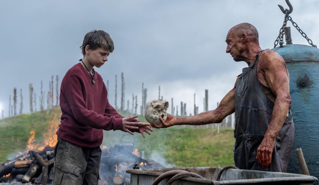 A man hands a boy a skull. They stand in front of a fire A man hands a boy a skull. They stand in front of a fire