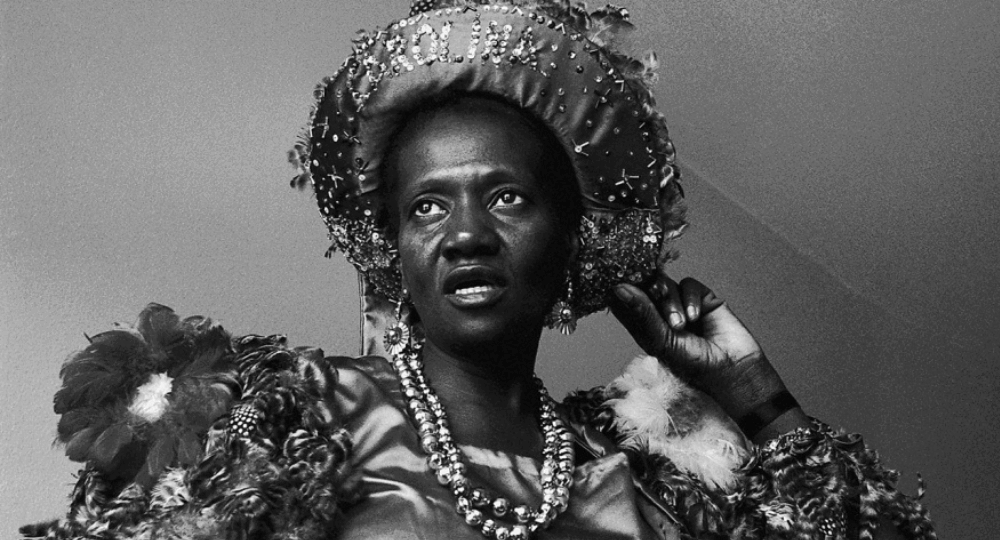 #descrevemospravocê  Fotografia em preto e branco retrata uma mulher negra vestida com trajes festivos e tradicionais. Ela usa um chapéu redondo e ornamentado, com bordados, miçangas, flores e franjas ao redor. Na parte frontal do chapéu, está bordado o nome "Carolina" em letras maiúsculas. A mulher olha levemente para cima e à esquerda, com expressão firme e altiva. Ela segura uma das mãos próximas ao rosto, com os dedos arqueados. Veste uma roupa decorada com tecido brilhante, flores, penas e rendas. No pescoço, colares de contas completam o figurino. A composição, com fundo neutro e iluminação suave, destaca os detalhes do rosto e dos adornos. A imagem transmite força, dignidade e uma presença marcante, evocando ancestralidade e identidade cultural.