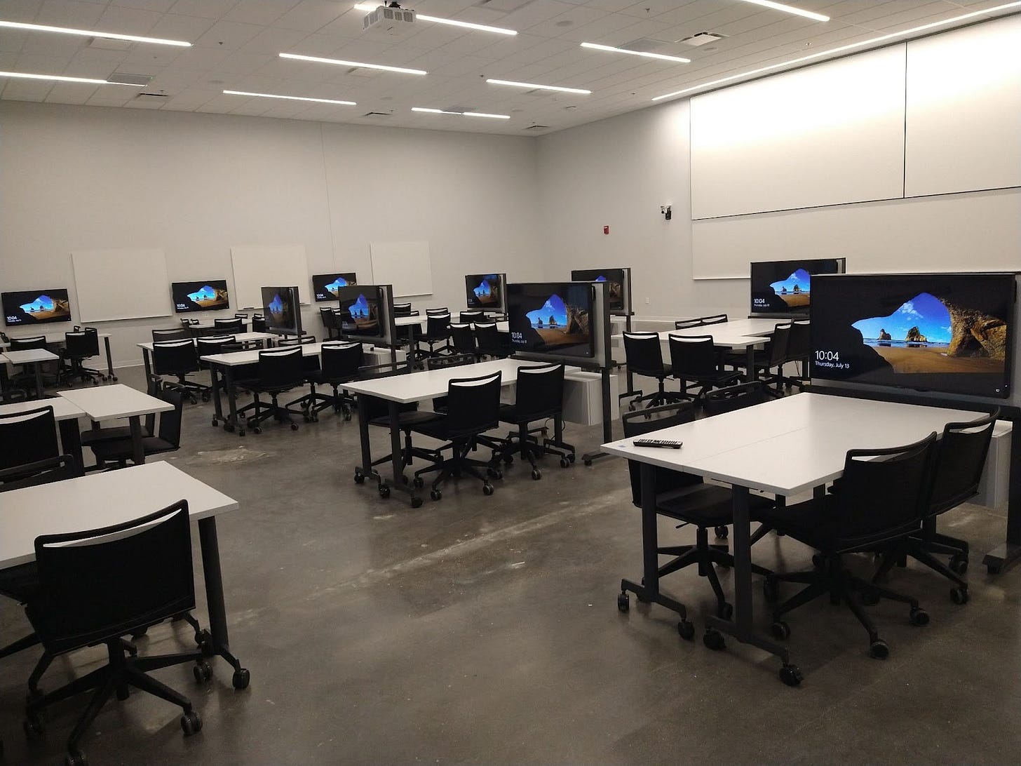 TEAL classroom with a high ceiling, two screens above a long whiteboard are along one wall. Along the other wall are smaller whiteboards and wall mounted displays. Chairs are arranged with tables in groups of four to six and the groups in the middle of the room are next to a digital display on a stand.