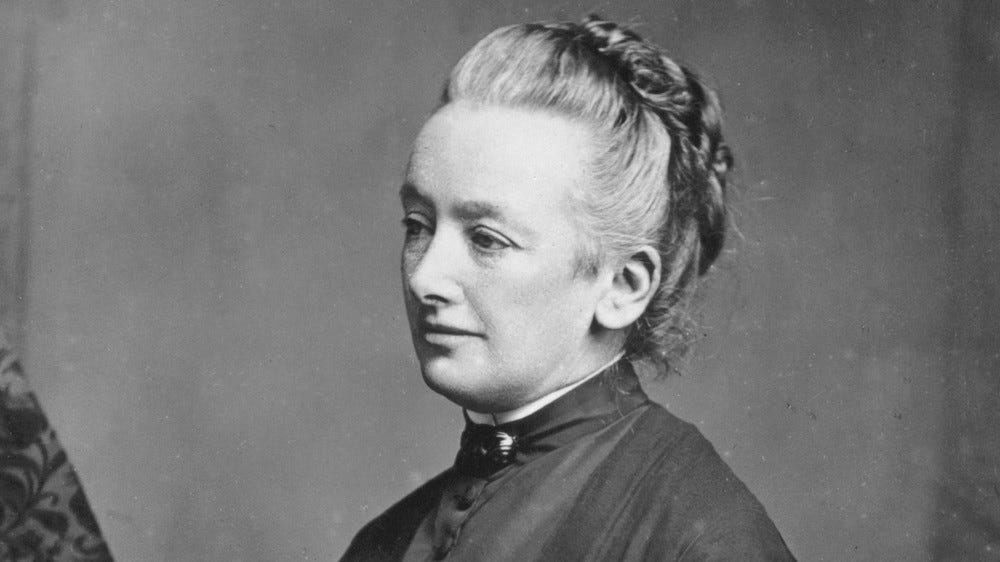 b/w head and shoulders shot of a woman looking off to the left of the image. Her hair is drawn back and up, and tied at the back of her head. She looks thoughtful
