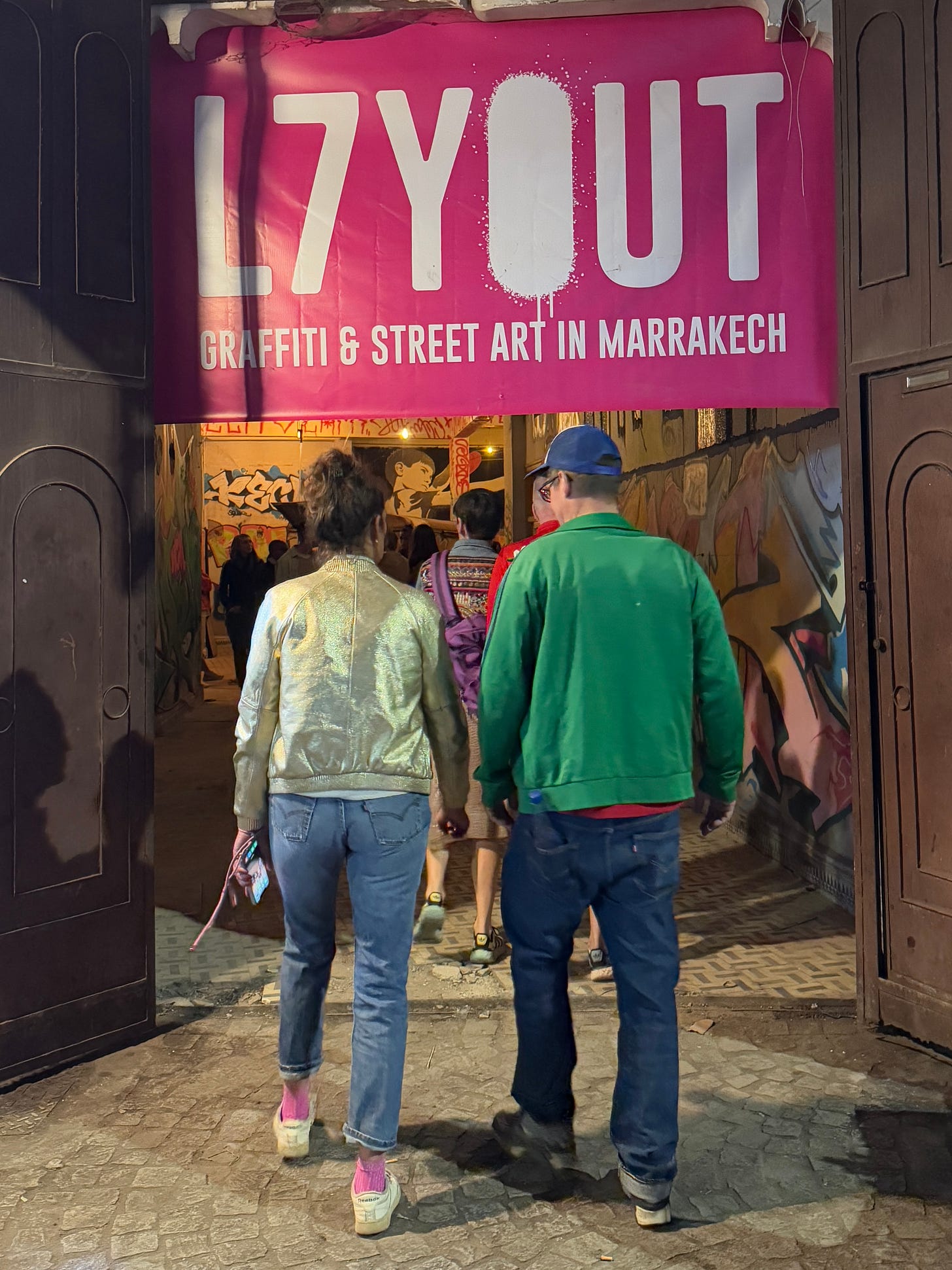 Visitors enter the L7YOUT street art gallery in Marrakech during the book release. A pink banner above the entrance reads “L7YOUT – Graffiti & Street Art in Marrakech.” The narrow passage inside is covered in graffiti and murals, with people walking in from the street, blending gallery space and everyday city life. Visitors enter the L7YOUT street art gallery in Marrakech during the book release. A pink banner above the entrance reads “L7YOUT – Graffiti & Street Art in Marrakech.” The narrow passage inside is covered in graffiti and murals, with people walking in from the street, blending gallery space and everyday city life.