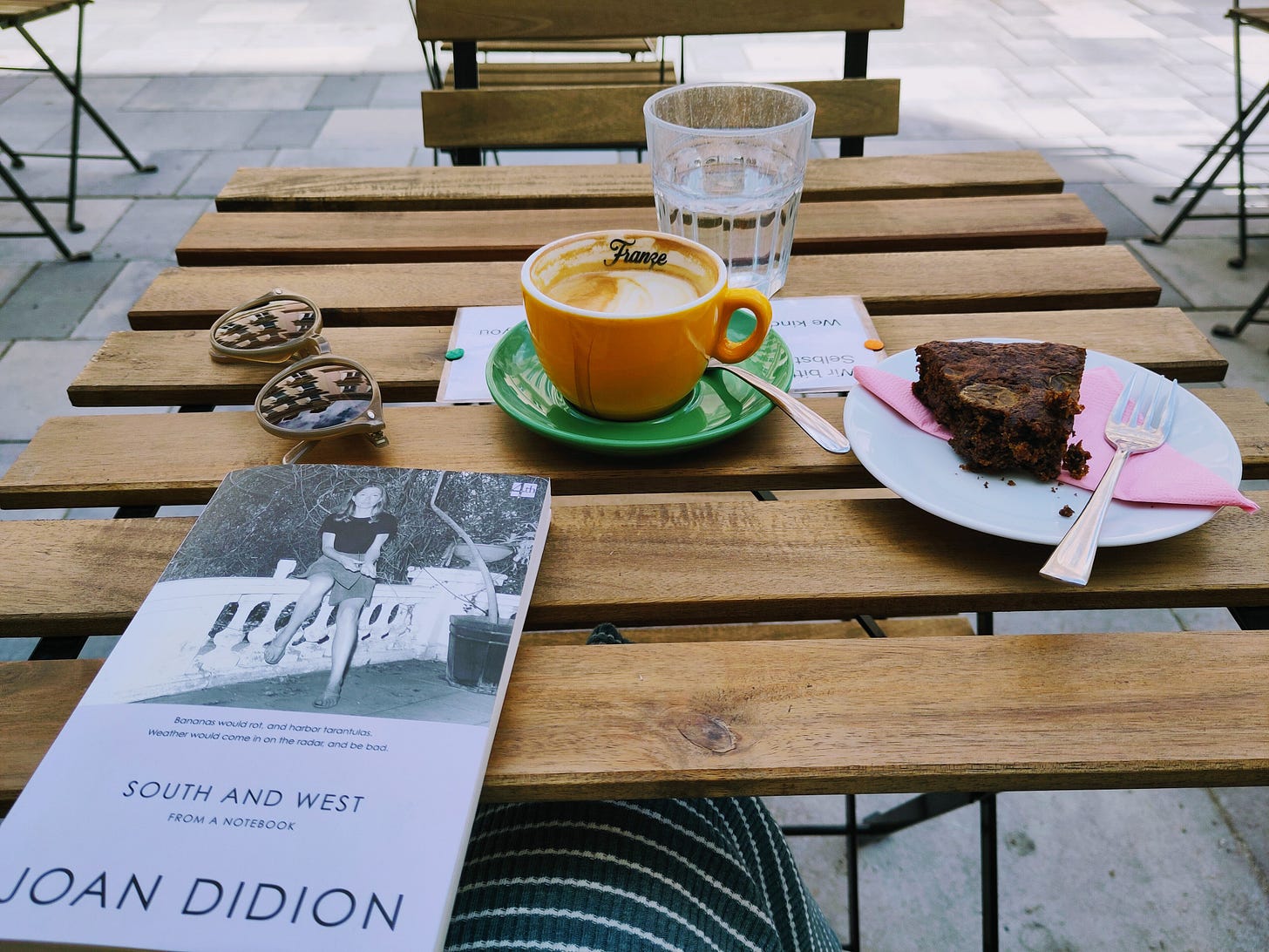 The photo shows a table at an outdoor space of a coffee shop. There's a cup of coffee, a piece of cake, a glass of water, sunglasses, and a copy of Joan Didion's book South and West on the table.