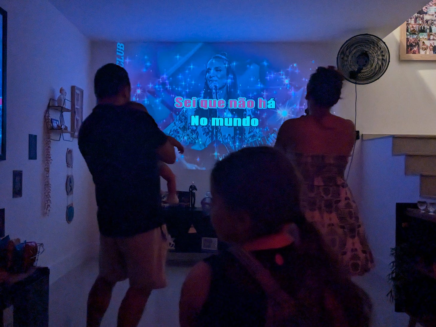 duas pessoas de costas cantando "a lua q eu t dei" da ivete sangalo no karaokê. a da esquerda é um homem segurando um bebê pequeno no colo, dele visível apenas o bracinho e a perna direita, e a pessoa da direita é uma mulher com o cabelo amarrado em coque e um vestido estampado. na frente deles na foto, está uma criança sentada, também de costas, olhando para o telão em que está uma umagem da ivete e a letra da canção