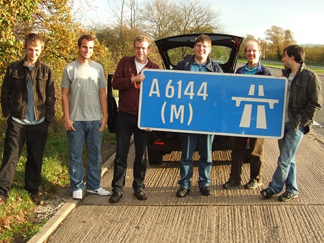Three photographs showing people posing with the A6144(M) sign in various settings - on the left in front of a building; in the middle, two people obscured by the signs and clearly having trouble holding them; on the right a whole group posing together with the sign in a lay-by
