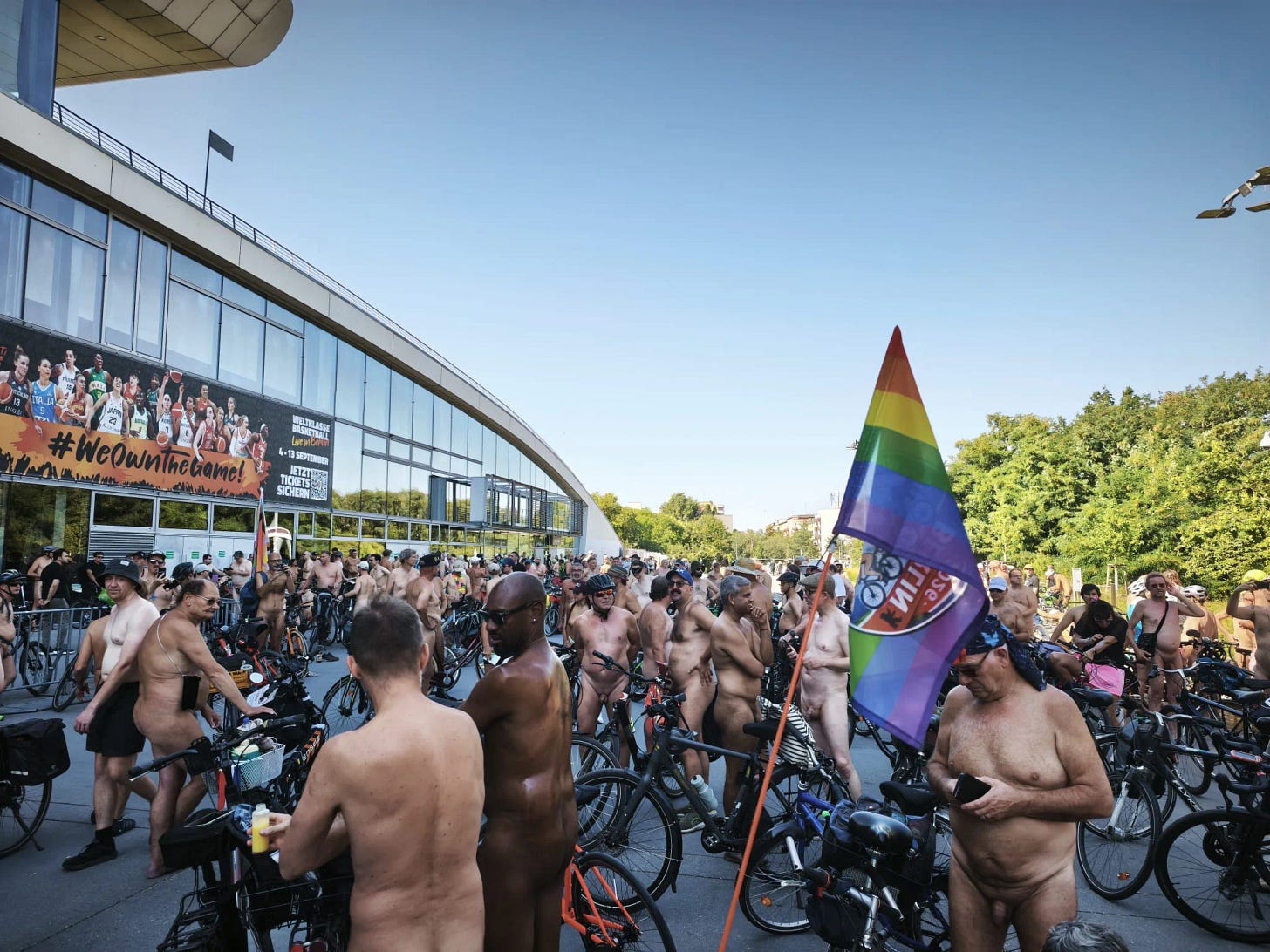 A large crowd of nude cyclists gather with their bicycles outside a modern building in Berlin during the World Naked Bike Ride. A rainbow flag rises above the group. A large crowd of nude cyclists gather with their bicycles outside a modern building in Berlin during the World Naked Bike Ride. A rainbow flag rises above the group.