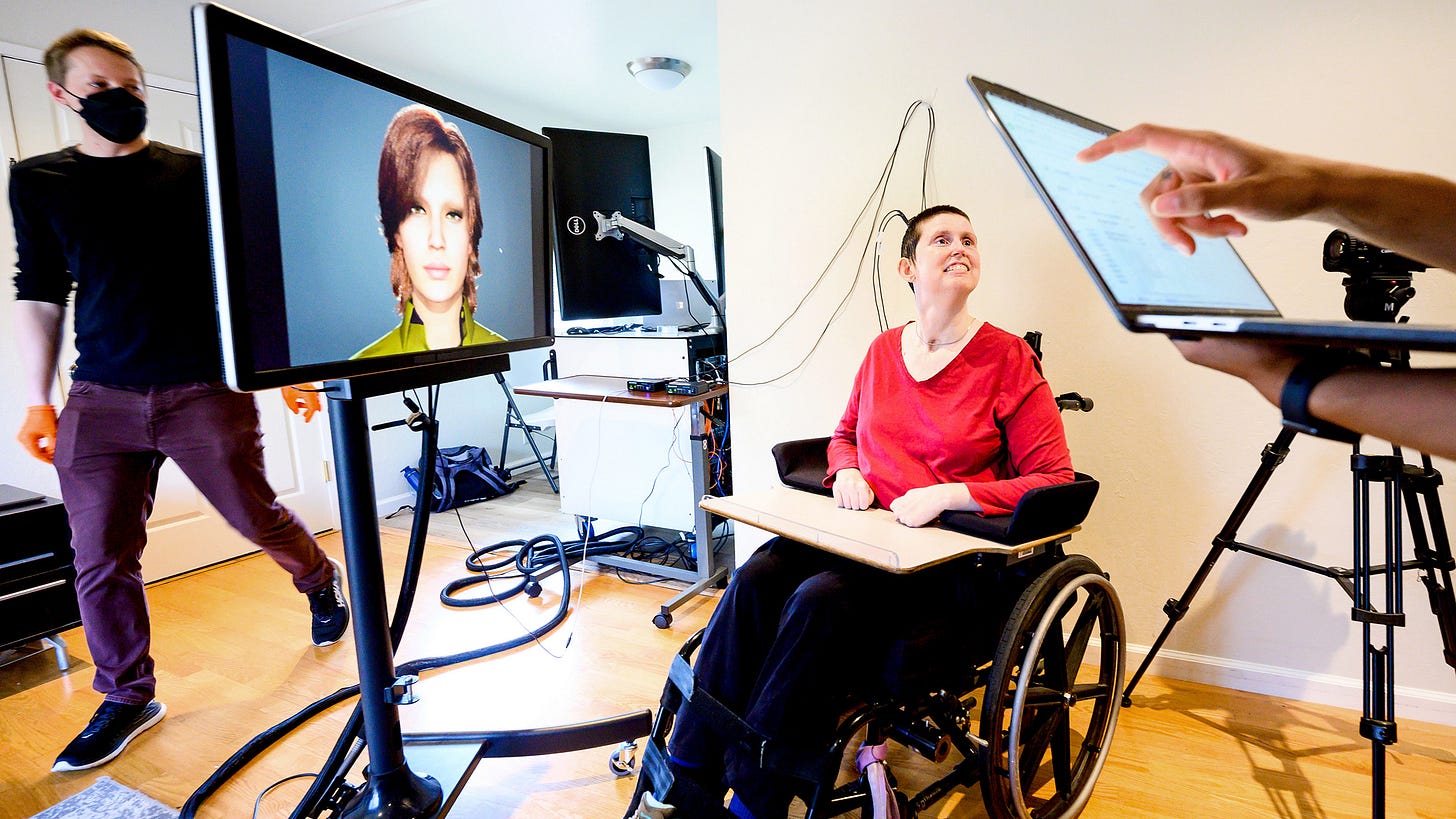 Research participant, sitting in a wheelchair, is connected by wires to a computer and monitor that displays an avatar. A research coordinator can be seen walking behind the monitor, and in the foreground, someone is holding a laptop computer. Research participant, sitting in a wheelchair, is connected by wires to a computer and monitor that displays an avatar. A research coordinator can be seen walking behind the monitor, and in the foreground, someone is holding a laptop computer.