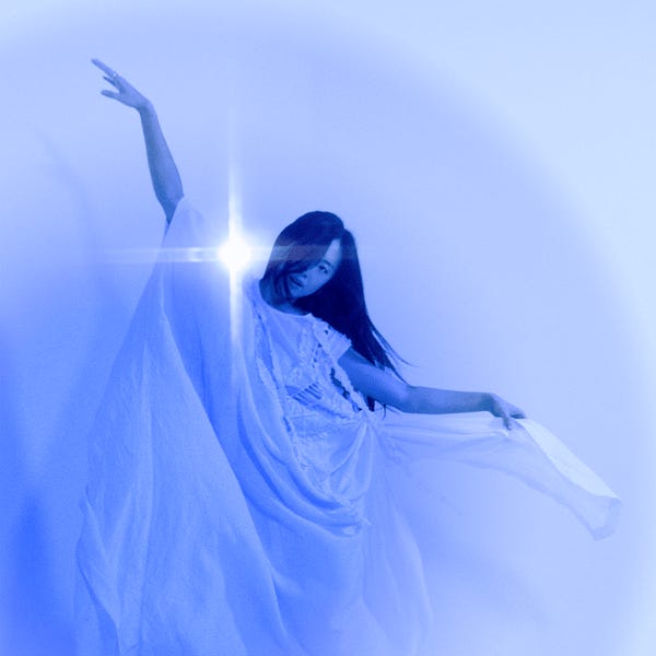 Sarah Kinsley stands in an etheral room, wearing a white gown, with her arms raised above her, the fabric of her dress draping down.