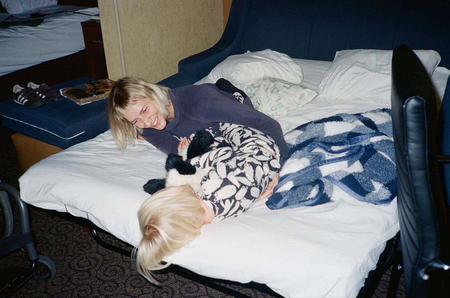 Rebekah is playing with her son on a fold-out bed covered in blankets and pillows. She leans over him, smiling. Her arms are around him. He's facing away from the camera, holding a lovie in his arms.