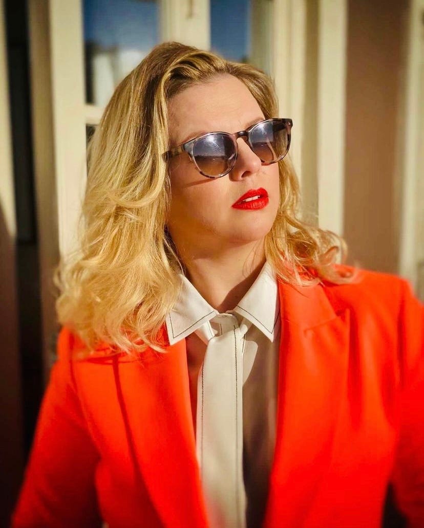 Amber poses outside, wearing sunglasses and a bright orange suit. Amber poses outside, wearing sunglasses and a bright orange suit.