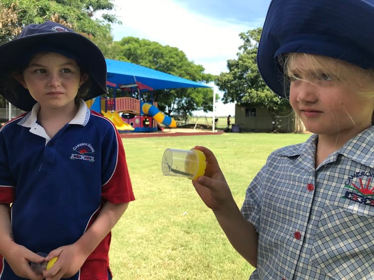 Two children in school uniforms hold up a specimen jar with an insect inside. Two children in school uniforms hold up a specimen jar with an insect inside.