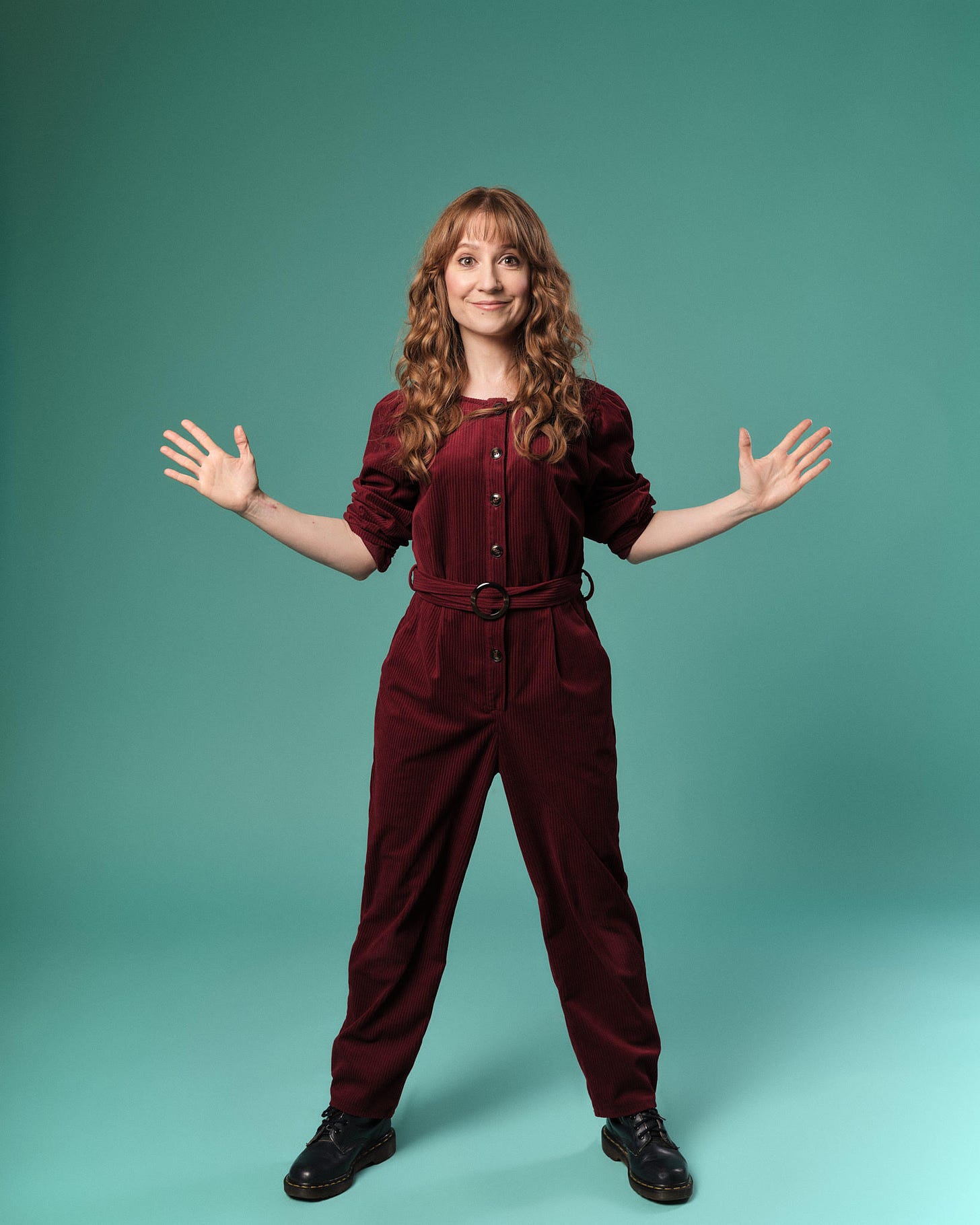 Woman in burgundy jumpsuit stands with her hands out and her legs in a wide stance. She has long ginger wavy hair