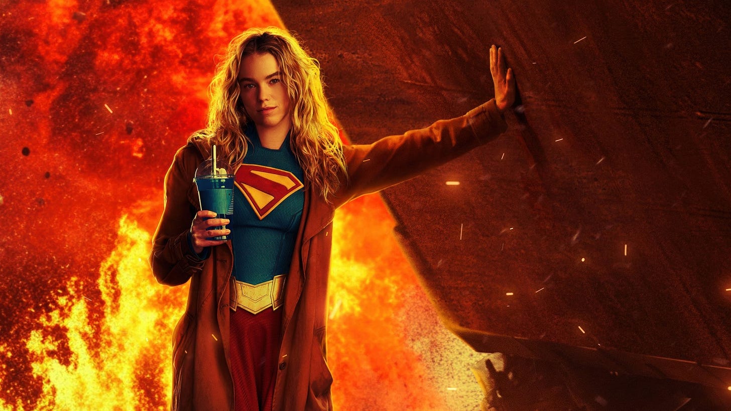 A imagem apresenta a atriz Milly Alcock caracterizada como Supergirl, posicionada de forma imponente no centro da composição diante de um cenário de pura destruição. Com cabelos loiros, longos e ondulados, ela encara a câmera com uma expressão firme e séria, vestindo o traje azul clássico com o símbolo do "S" estilizado no peito e uma capa vermelha que se estende por trás de seus ombros. Em uma postura que mistura força e casualidade, ela estende o braço esquerdo para apoiar a mão espalmada contra um grande fragmento de metal escuro, enquanto segura com a mão direita um copo azul com tampa e canudo. O fundo é dominado por uma explosão massiva, preenchido por chamas intensas em tons de laranja e vermelho, além de fagulhas e poeira flutuando pelo ar, o que acentua a atmosfera de pós-batalha da cena.