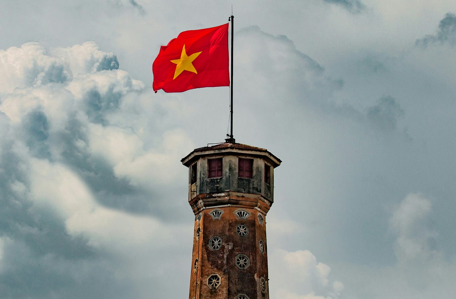 The red Vietnamese flag with a yellow star in the center flies on a pole, above a lighthouse. The red Vietnamese flag with a yellow star in the center flies on a pole, above a lighthouse.