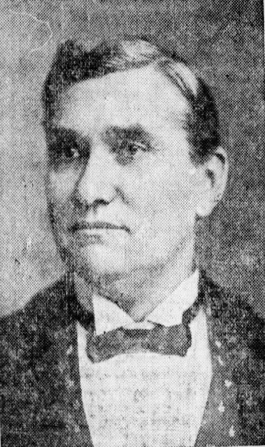 A black-and-white photograph of a man dressed in a smart suit and a shirt with wing-tip collar and a bow tie. He is middle-aged, white, clean-shaven and has short neatly cut hair. He has a neutral expression and is looking slightly to his right.