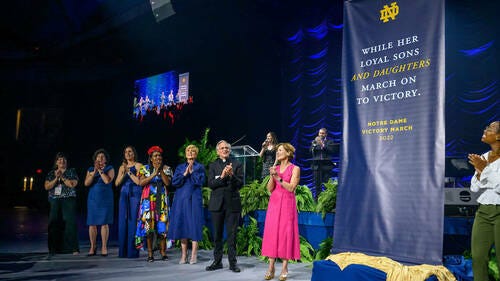 Fr. John Jenkins stands among women graduates of the University of Notre Dame a a banner is unveiled reading "While her loyal sons and daughters march on to victory"