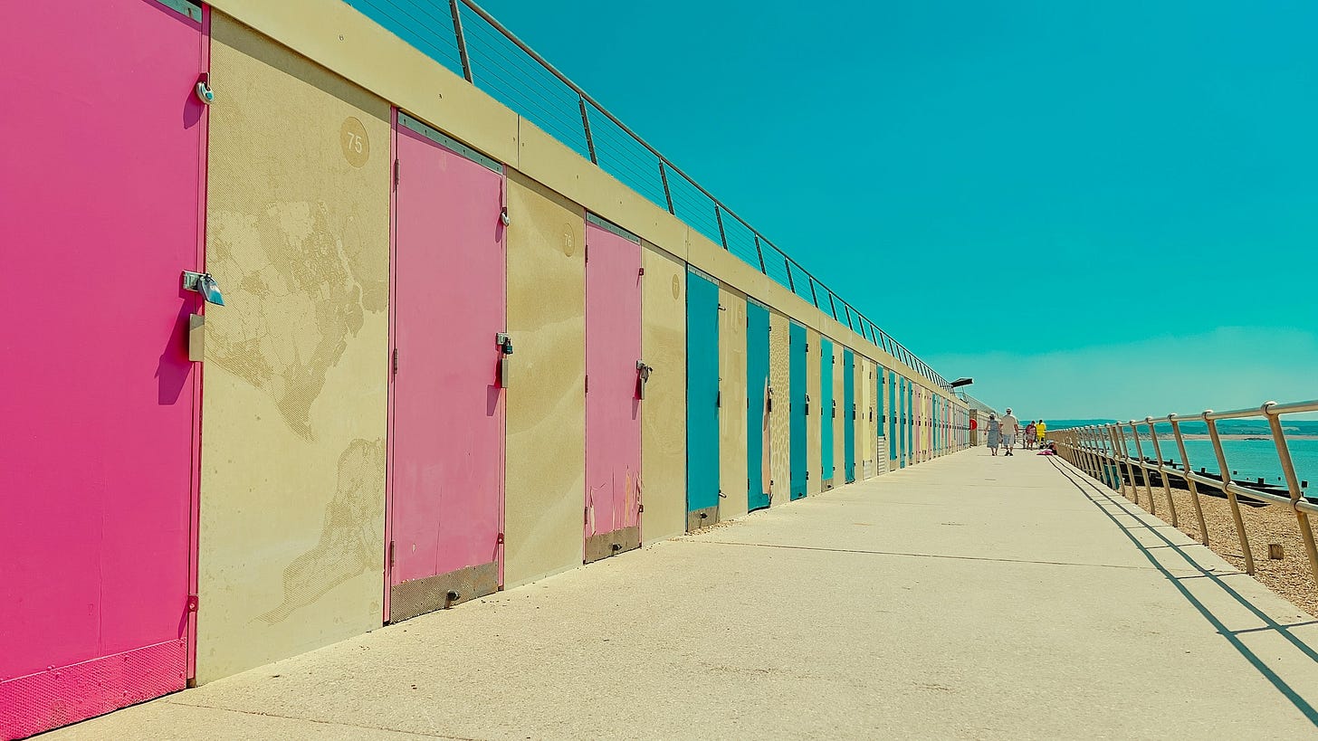 Cabine da spiaggia con porte rosa e turchese, allineate lungo una passerella di cemento. Le cabine, dalla facciata beige, si estendono verso l'orizzonte sotto un cielo azzurro e limpido. La ringhiera di metallo che delimita il bordo della passerella è visibile a destra, con l'oceano e la spiaggia sottostante. Sullo sfondo, si intravedono alcune persone che passeggiano.