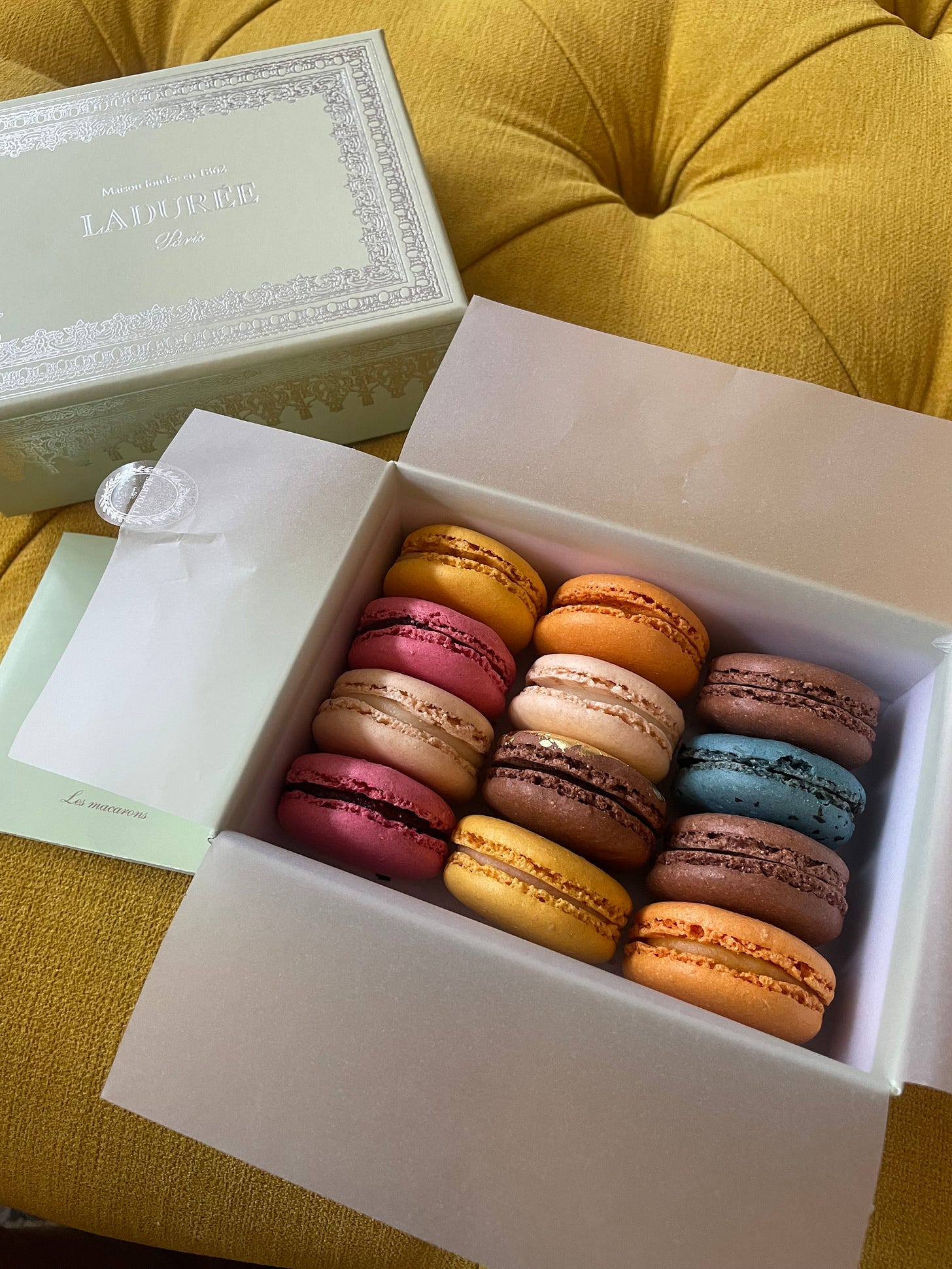 An open box of Laduree macarons placed on a yellow piece of furniture. The box contains 12 macarons of different colours and flavours nestled in delicate tissue paper.