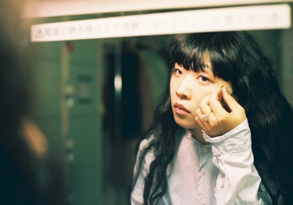 A photo of Ichiko Aoba's reflection in a mirror. She is applying make-up to her face. A photo of Ichiko Aoba's reflection in a mirror. She is applying make-up to her face.