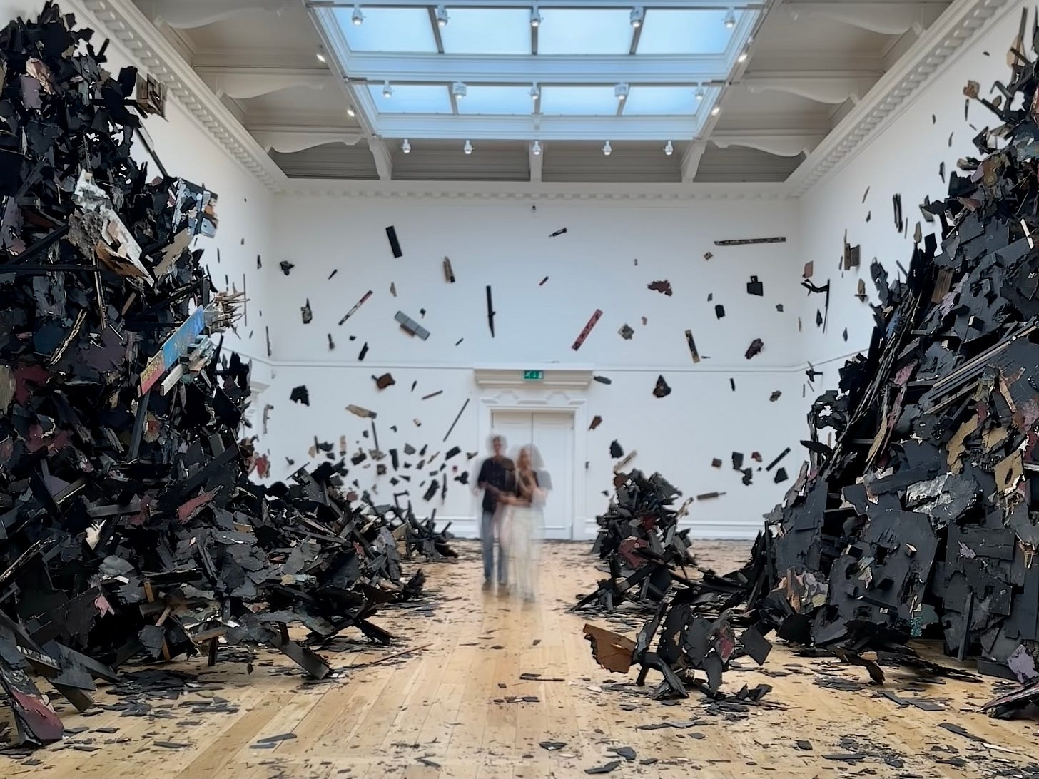 An art installation in a gallery that looks like an explosion of blackened matter, caught in the moment, with fragments frozen in space. Two figures are out of focus in the middle as part of this long exposure shot.