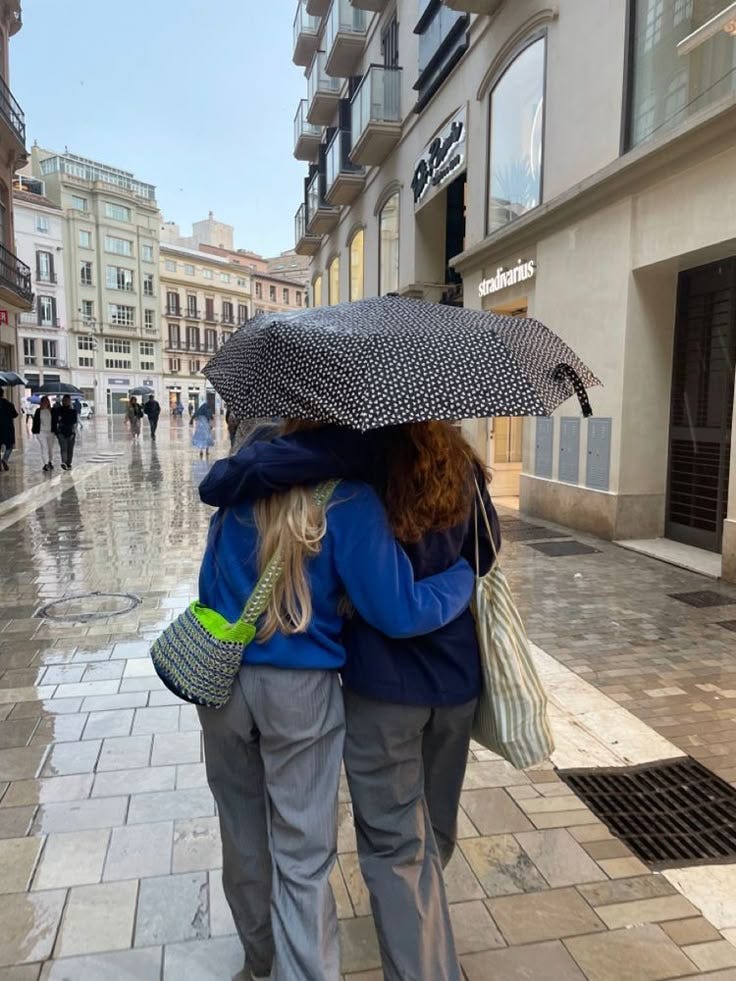 This may contain: two women walking down the street with an umbrella over their heads and bags on their backs