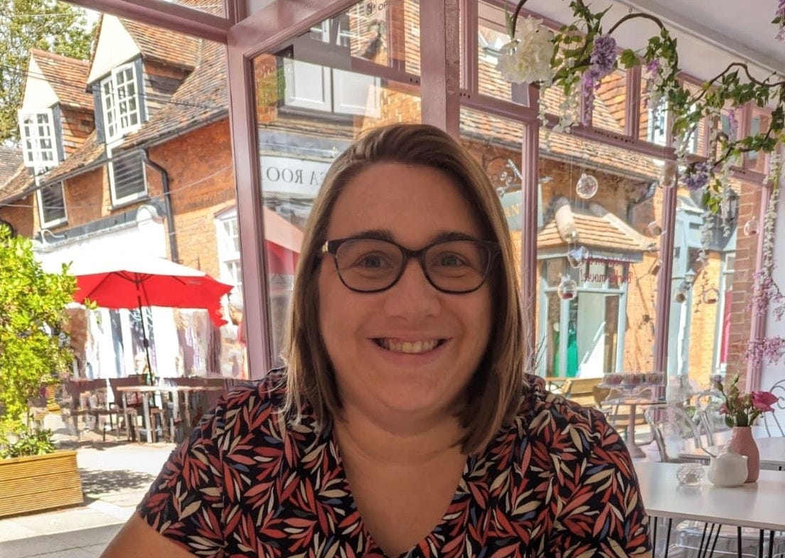 Photograph of Sian Shaw. A white woman in her 30s with straight blonde/brown hair wearing dark glasses and a colourful dress. She is smiling. The background shows an empty café.