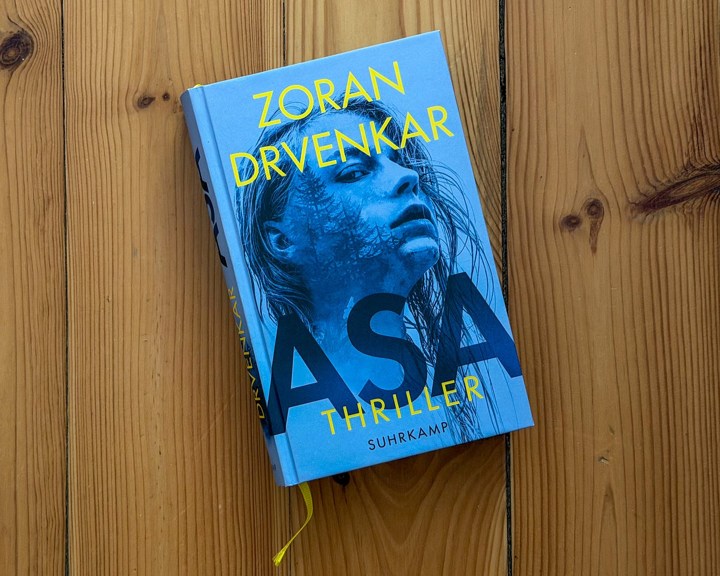 Buchcover des Thrillers „Asa" von Zoran Drvenkar, erschienen im Suhrkamp Verlag. Das Hardcover liegt auf einem Fußboden aus hellem Kiefernholz. Der Einband ist in einem intensiven Blauton gehalten und zeigt das Gesicht einer jungen Frau, deren nasse Haare ihr ins Gesicht fallen. In ihr Gesicht ist wie eine Doppelbelichtung eine dunkle Baumsilhouette eingearbeitet. Ihr Blick ist nach oben gerichtet. Der Autorenname „Zoran Drvenkar" steht in Großbuchstaben in leuchtendem Gelb oben auf dem Cover, der Titel „ASA" in großen dunkelblauen Versalien in der Bildmitte. Darunter stehen in kleineren gelben Buchstaben „Thriller" und „Suhrkamp". Am unteren Buchrand ist ein gelbes Lesebändchen zu sehen. Buchcover des Thrillers „Asa" von Zoran Drvenkar, erschienen im Suhrkamp Verlag. Das Hardcover liegt auf einem Fußboden aus hellem Kiefernholz. Der Einband ist in einem intensiven Blauton gehalten und zeigt das Gesicht einer jungen Frau, deren nasse Haare ihr ins Gesicht fallen. In ihr Gesicht ist wie eine Doppelbelichtung eine dunkle Baumsilhouette eingearbeitet. Ihr Blick ist nach oben gerichtet. Der Autorenname „Zoran Drvenkar" steht in Großbuchstaben in leuchtendem Gelb oben auf dem Cover, der Titel „ASA" in großen dunkelblauen Versalien in der Bildmitte. Darunter stehen in kleineren gelben Buchstaben „Thriller" und „Suhrkamp". Am unteren Buchrand ist ein gelbes Lesebändchen zu sehen.
