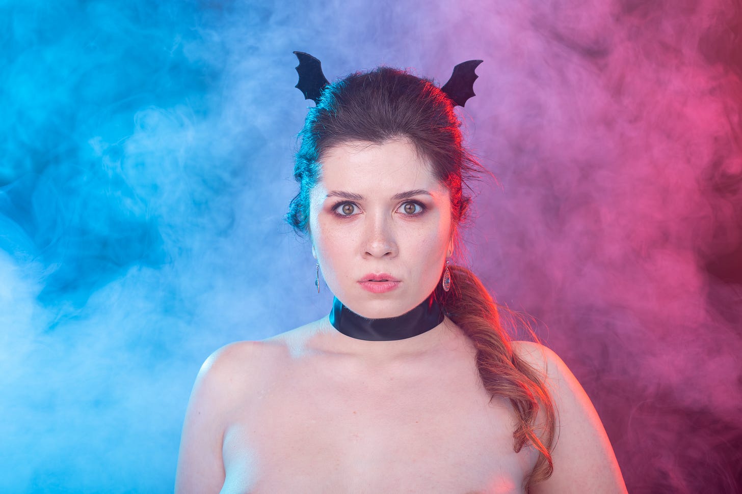 A woman wearing bat-shaped headband accessories and a black choker stands against smoky blue and pink lighting, evoking a playful Halloween atmosphere.