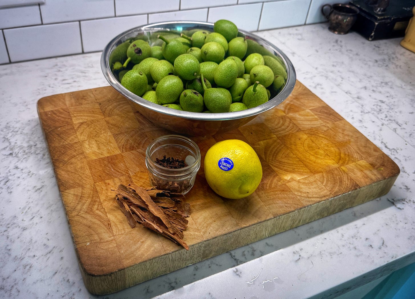 A large bowl of green walnuts sits on a cutting board. In front of it there is an open jar of cloves, a lemon, and a pile of canela strips A large bowl of green walnuts sits on a cutting board. In front of it there is an open jar of cloves, a lemon, and a pile of canela strips