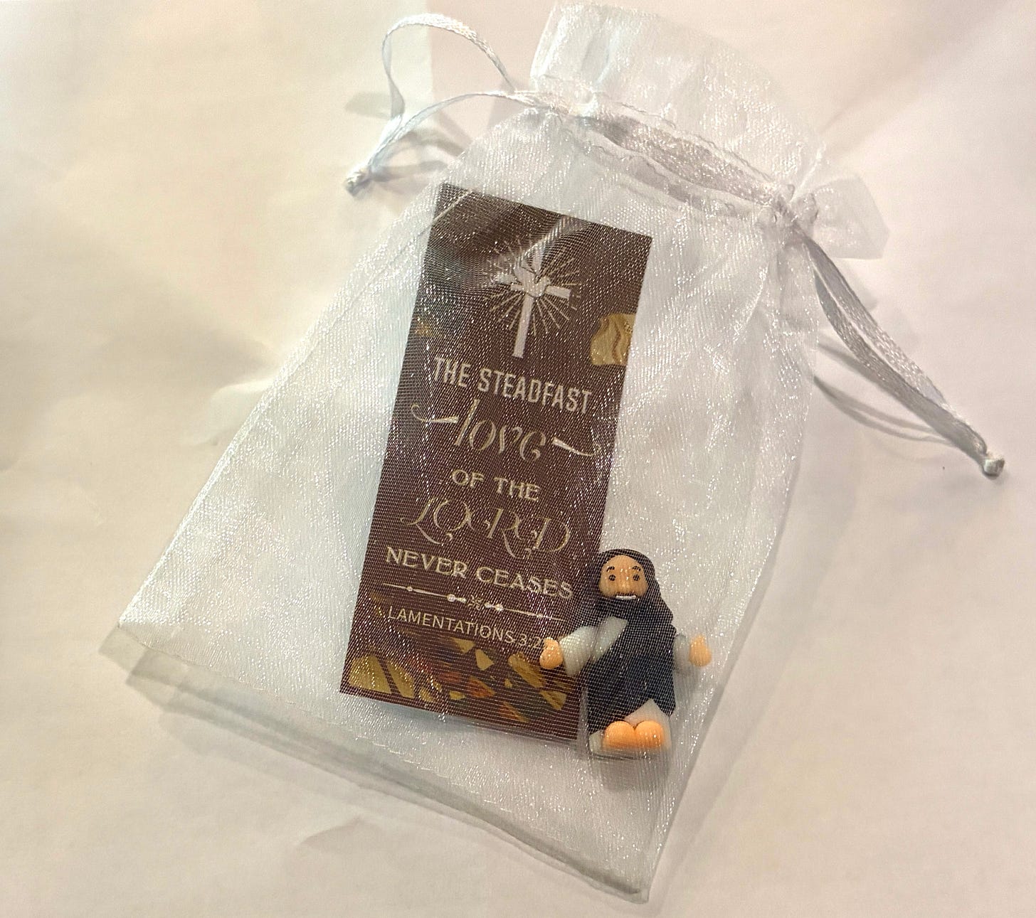 Small gift from Dosser Garage featuring a bookmark with the scripture “The steadfast love of the Lord never ceases” from Lamentations 3:22 and a tiny Jesus figurine tucked inside a sheer white drawstring bag.
