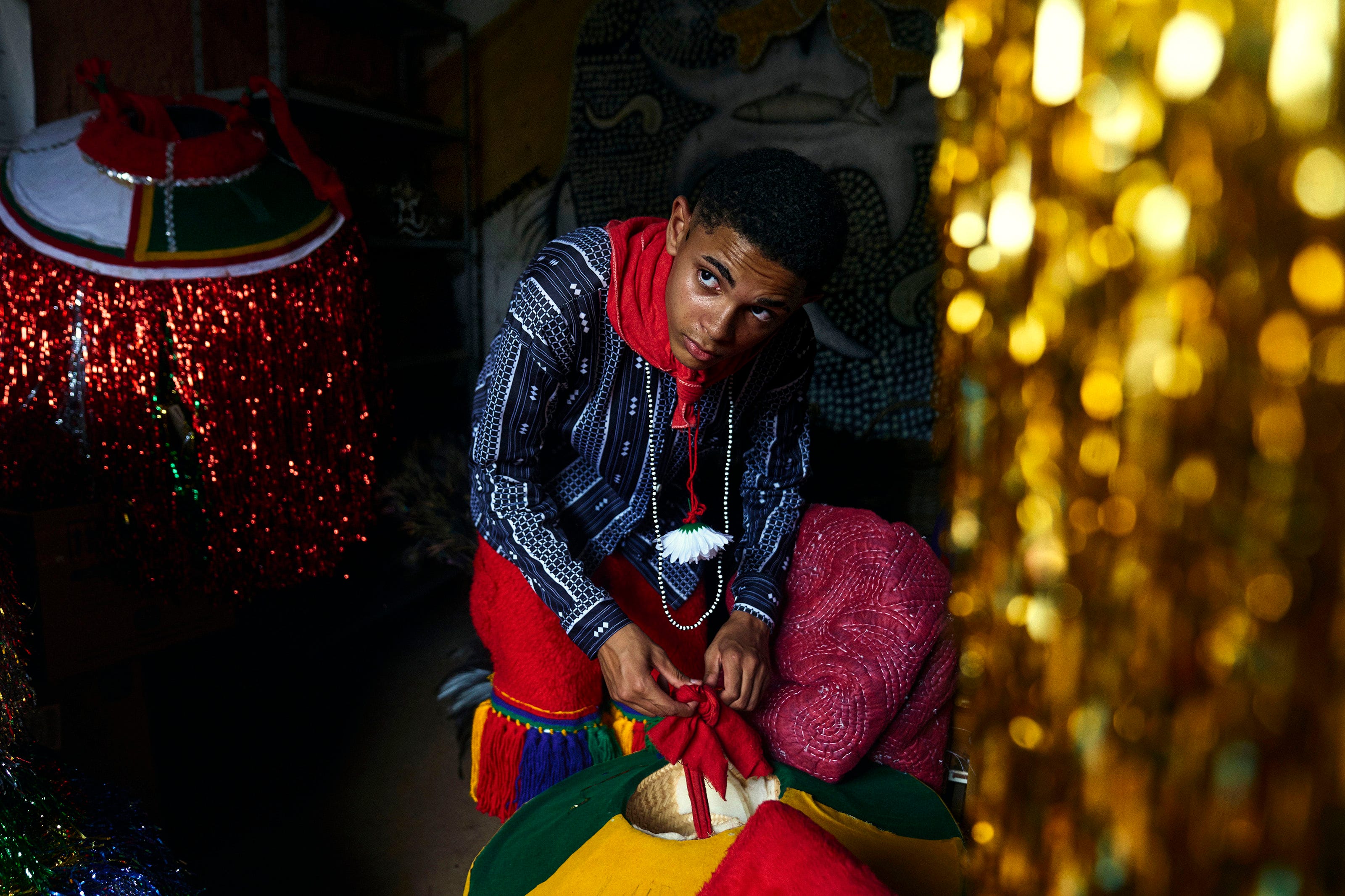 A person in colorful traditional clothing crouches and ties a red sash, surrounded by festive decorations with shiny tinsel and fabrics. They look upward, illuminated by soft, dramatic lighting.