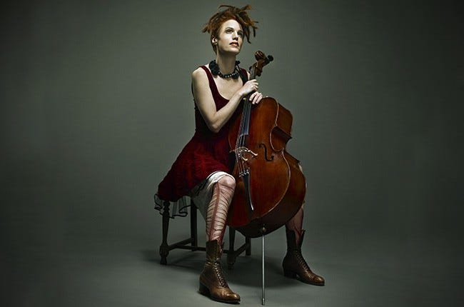 An image of a woman sitting on a stool in a dark grey room. She wears a deep red velvet dress, leather boots and torn red tights, a chunky black necklace, and has dark red dreadlocks. A cello sits between her knees