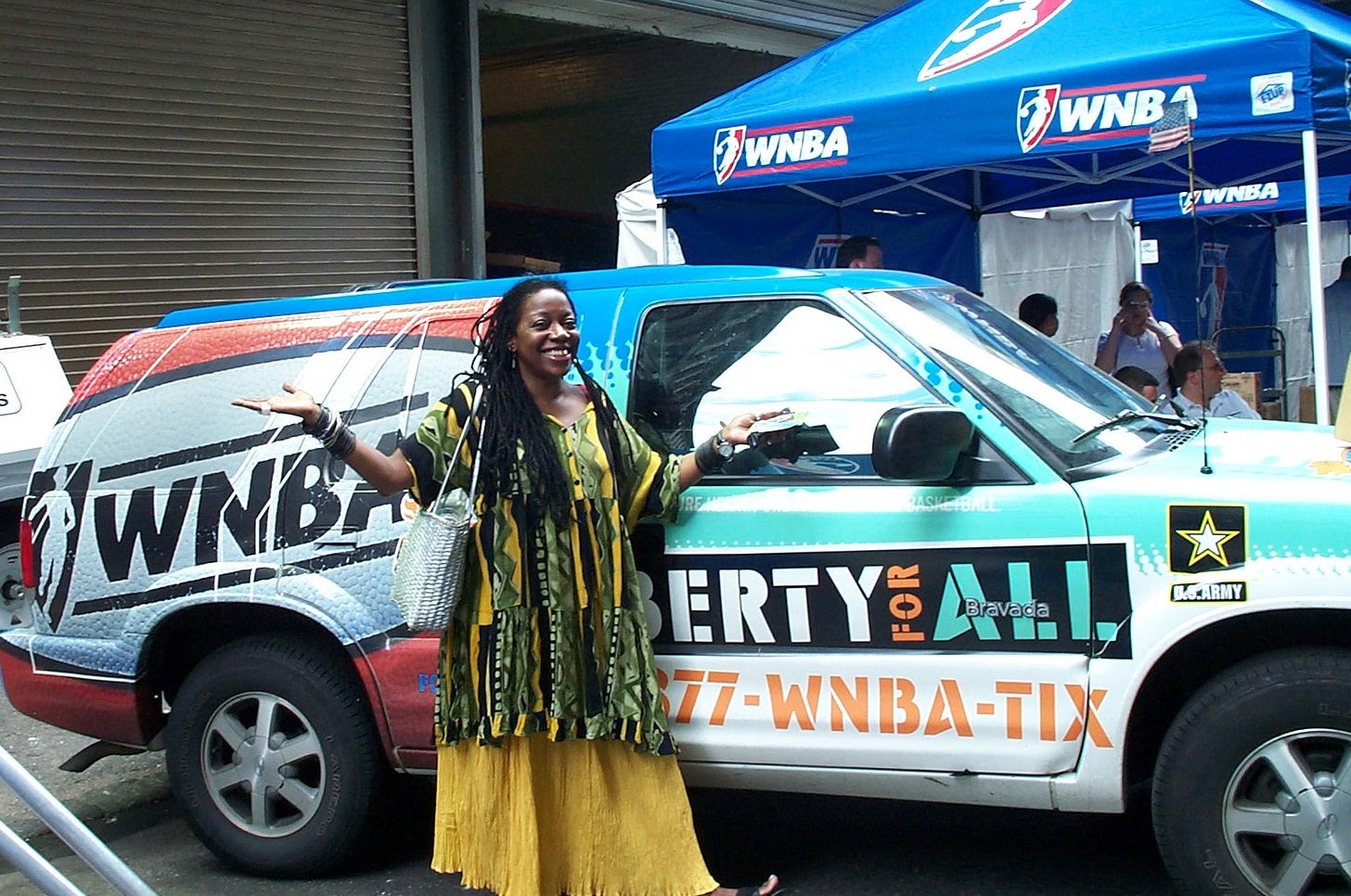 A woman of African descent with long locks dressed in a yellow and green flowing outfit, standing in front of a car painted with the words WNBA Liberty for All. 